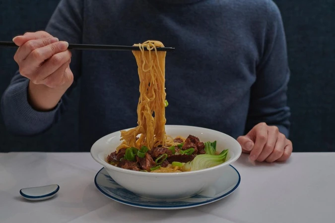 Close-up of a bowl of noodle soup with braised meat and greens, held up with chopsticks in a restaurant setting, food photography.