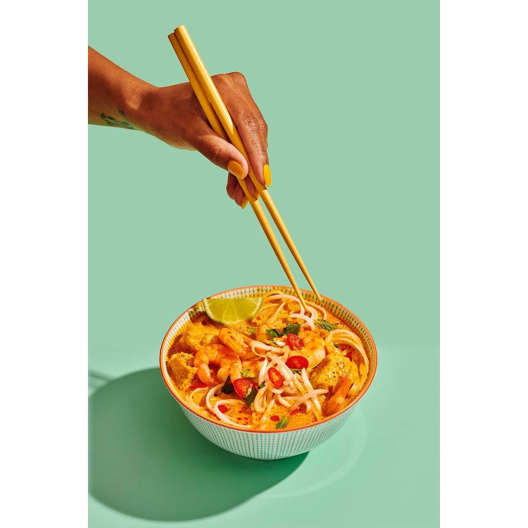 Overhead shot of hand using chopsticks to eat Asian noodle soup with shrimp, tofu, and lime, set against a vibrant mint-green backdrop.