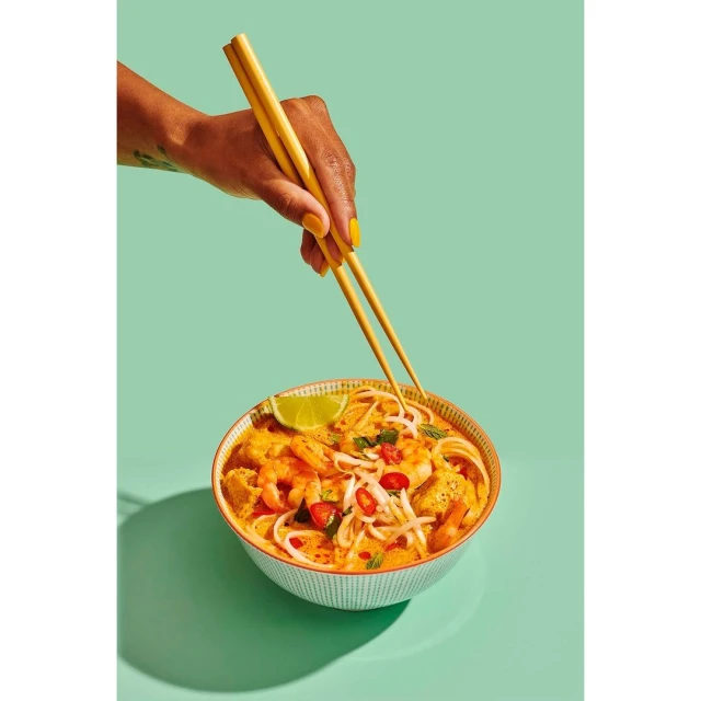 Overhead shot of hand using chopsticks to eat Asian noodle soup with shrimp, tofu, and lime, set against a vibrant mint-green backdrop.