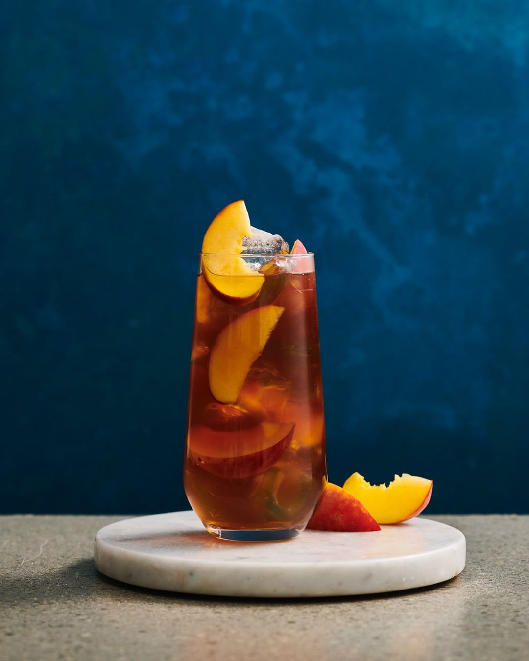 A clear glass of iced tea containing ice and peach slices, sitting on a marble stand against a dark blue background.