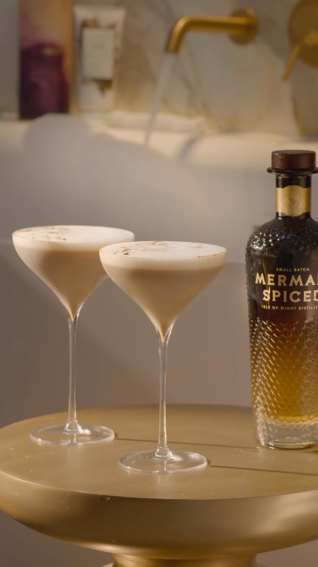 Two cocktails with a bottle of Mermaid Spiced vodka on a gold table in a bright bathroom with a bubble-filled bathtub in the background.