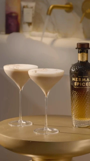 Two cocktails with a bottle of Mermaid Spiced vodka on a gold table in a bright bathroom with a bubble-filled bathtub in the background.
