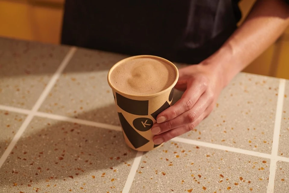 Close-up of a paper cup of cappuccino held in hand, on a patterned table with yellow wall in the background.