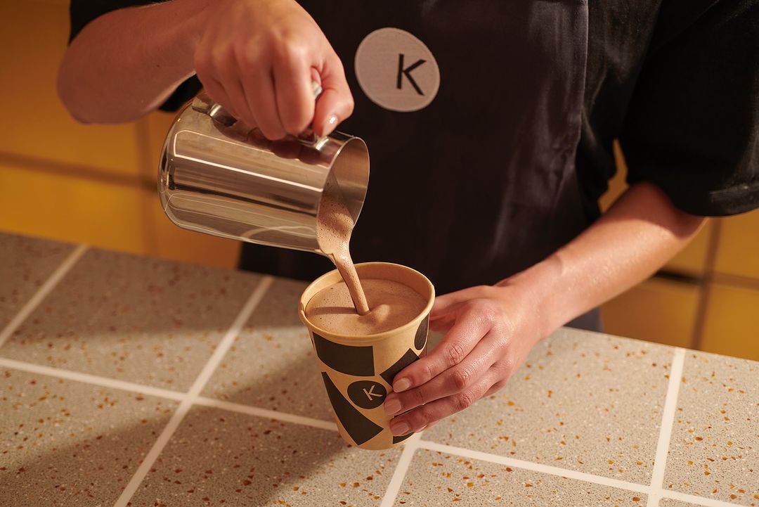 Close-up of a barista pouring a chocolate drink from a stainless steel pitcher into a branded paper cup on a tiled counter.