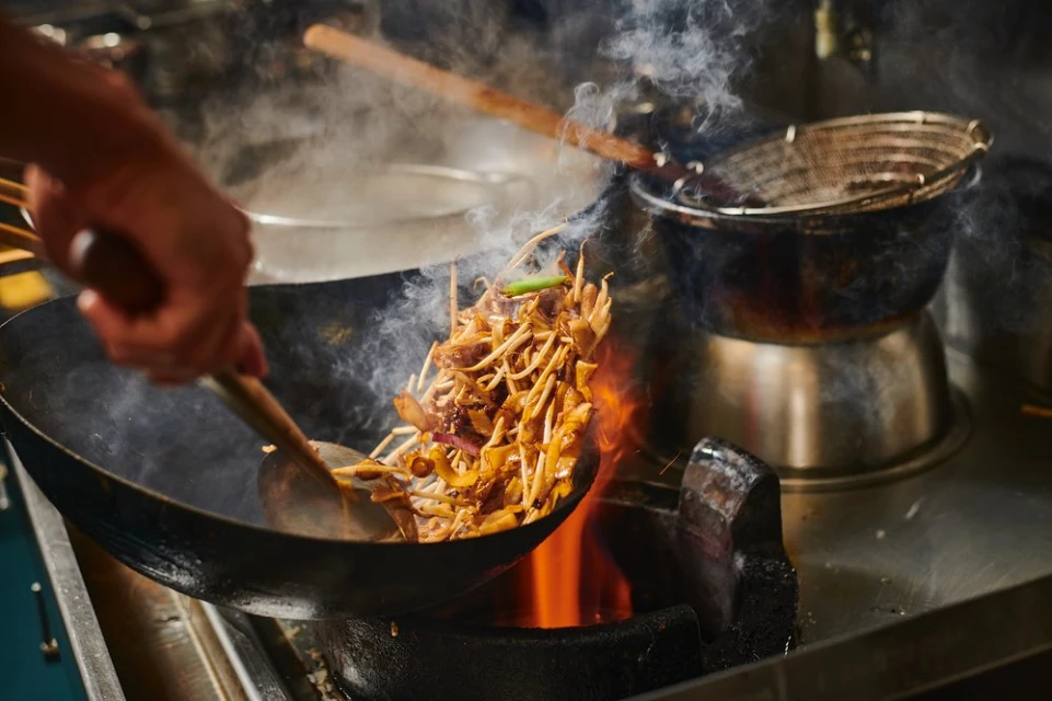 Cooking food in a wok on a stove with a wooden spatula, producing smoke and flames in a commercial kitchen setting.