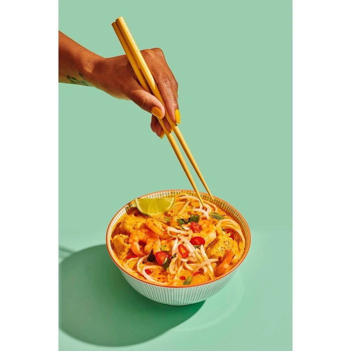 Close-up of a hand holding yellow chopsticks over a bowl of noodle soup with shrimp against a teal background.