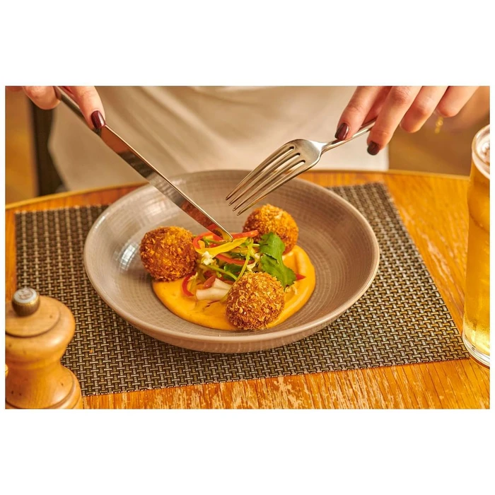 Close-up of hands using a knife and fork to cut golden fried arancini balls served on a vibrant orange puree with a fresh salad garnish.