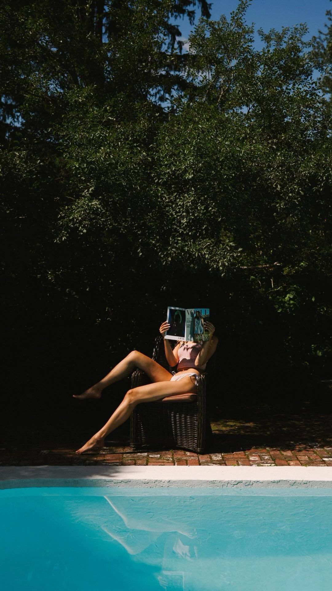 Woman in a bikini relaxes by the pool in an outdoor setting, reading a magazine in a chair, surrounded by lush greenery and trees.
