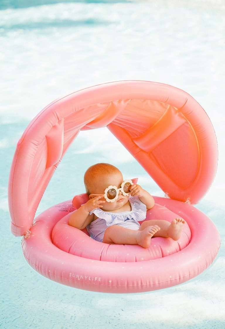 Infant in a pink inflatable pool float with a canopy, wearing flower-shaped sunglasses, relaxing in clear, shallow water on a sunny day.