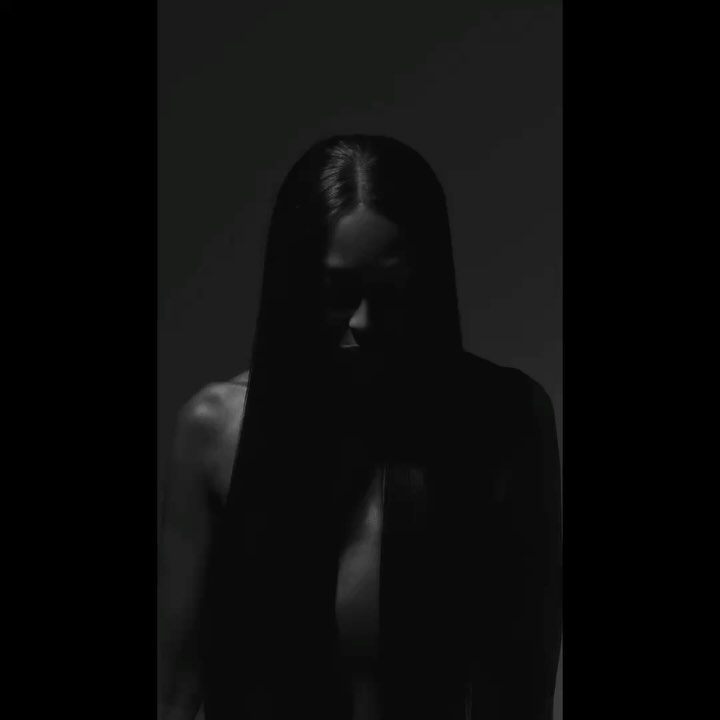 Monochrome portrait of an adult female with long dark hair and bare shoulders, set against a dark grey background in low light.