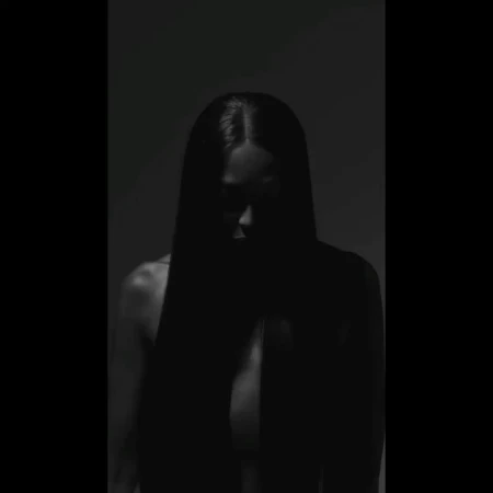 Monochrome portrait of an adult female with long dark hair and bare shoulders, set against a dark grey background in low light.