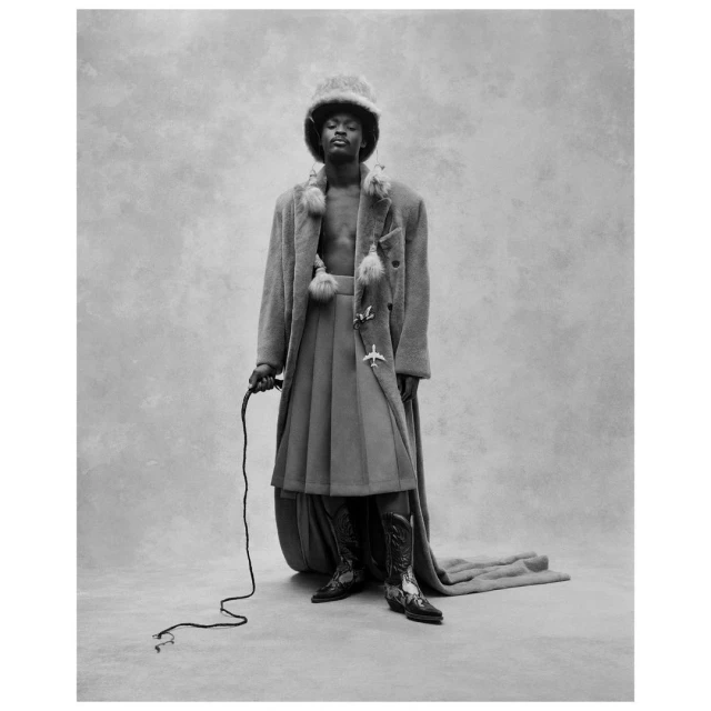 Monochromatic portrait of a male model in a fur hat, long coat, pleated skirt, and boots, holding a riding crop in a studio setting.