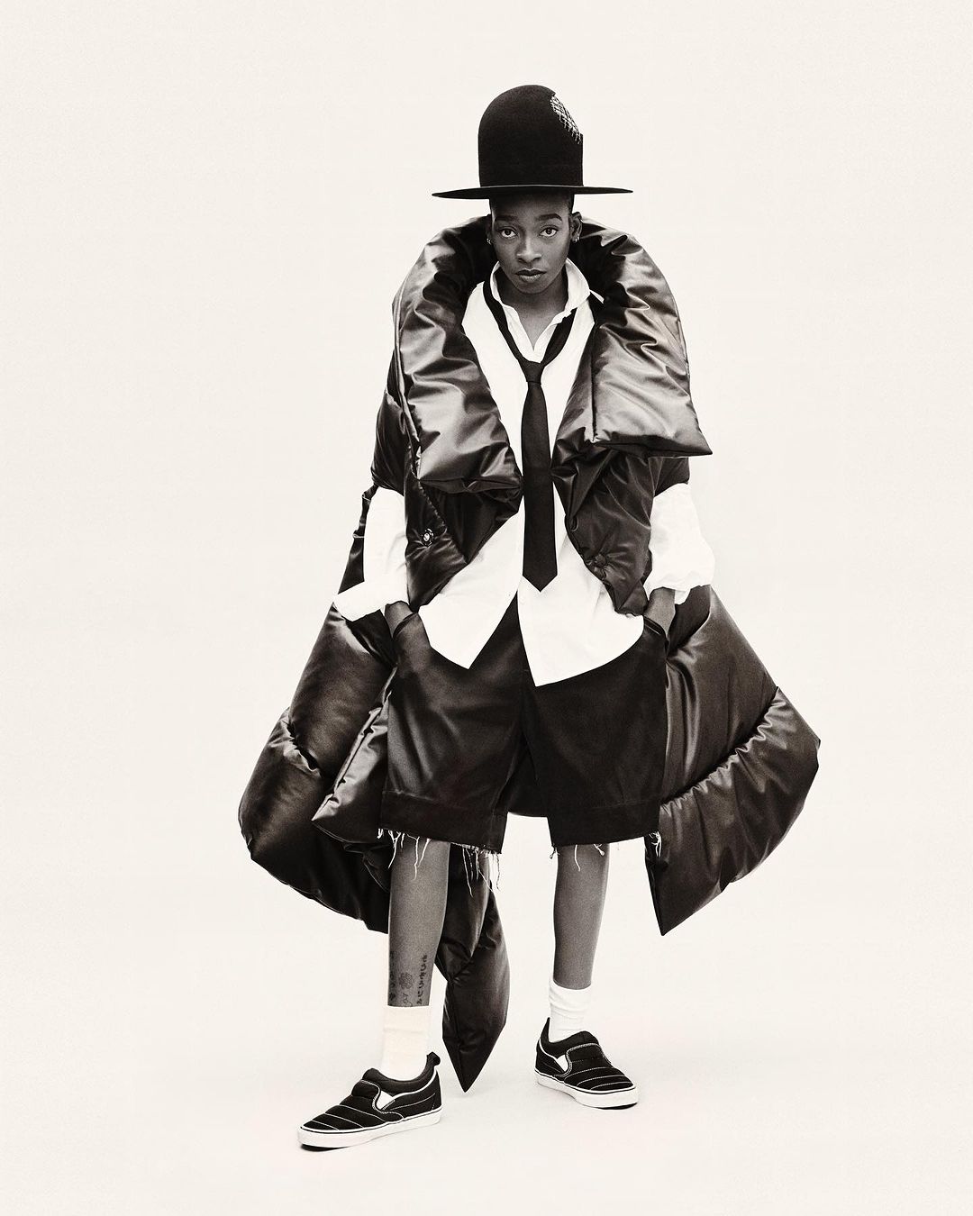 Fashion-forward individual in a large black puffer vest, white shirt, tie, shorts, hat, and sneakers, poses on a white backdrop in a studio shot.
