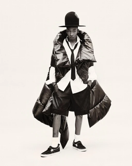 Fashion-forward individual in a large black puffer vest, white shirt, tie, shorts, hat, and sneakers, poses on a white backdrop in a studio shot.