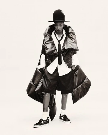 Fashion-forward individual in a large black puffer vest, white shirt, tie, shorts, hat, and sneakers, poses on a white backdrop in a studio shot.