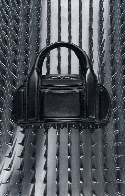 Black leather purse with studded trim displayed against a metallic background with vertical triangular spike details. Fashion accessory.