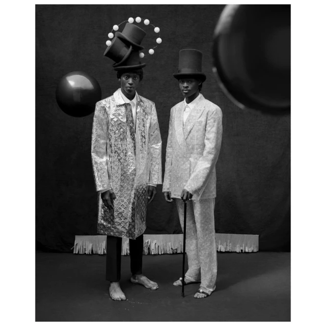 Monochrome fashion shot of two barefoot men in hats and ornate jackets, posing in a studio against a dark backdrop with props.