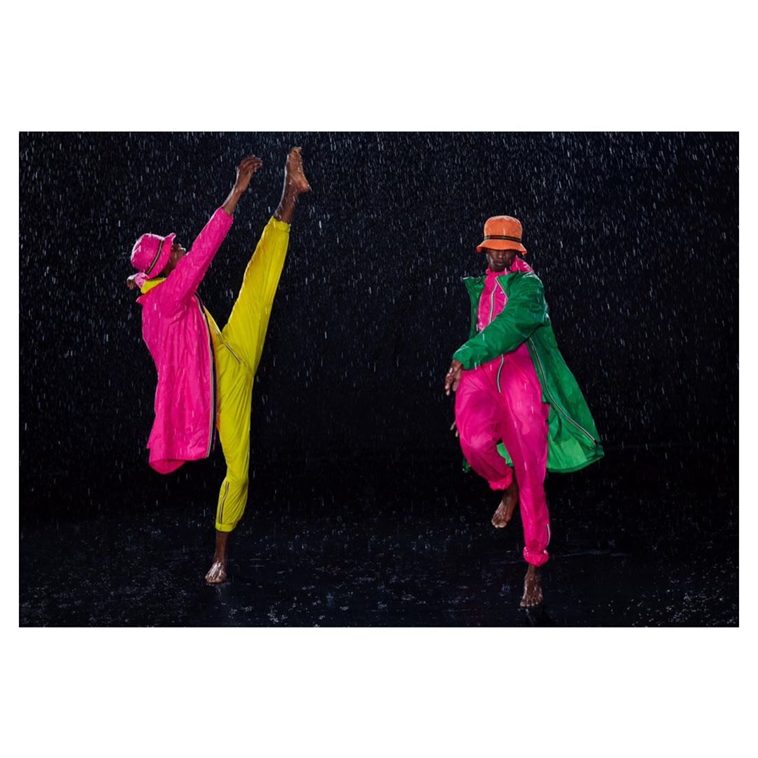 Two barefoot adult male fashion models pose in colorful rain gear against a dark backdrop with falling rain, showcasing dynamic movement.