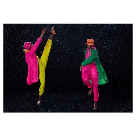 Two barefoot adult male fashion models pose in colorful rain gear against a dark backdrop with falling rain, showcasing dynamic movement.