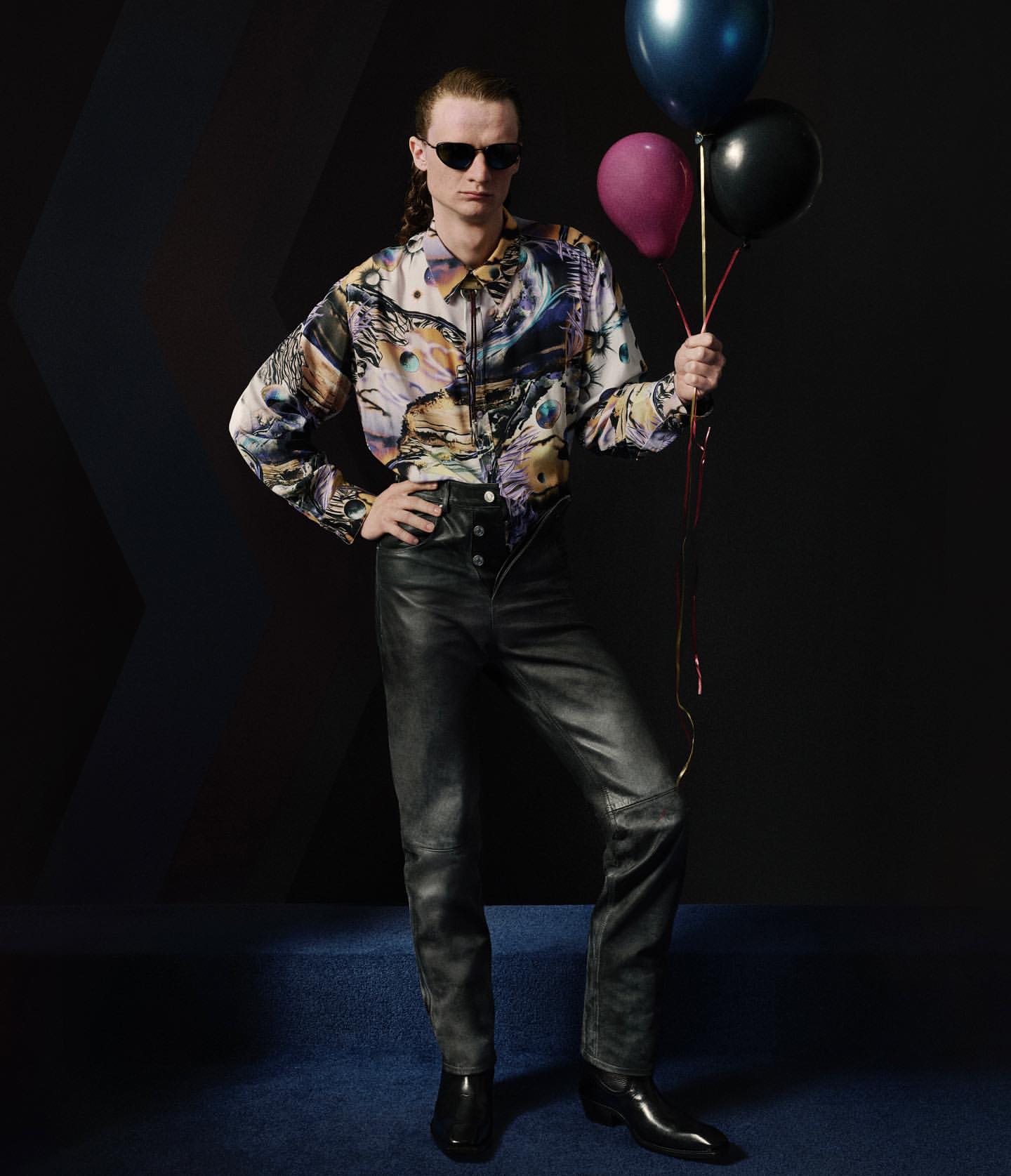 A male model with sunglasses poses in a studio, wearing a printed shirt and leather pants, holding black, blue, and pink balloons.