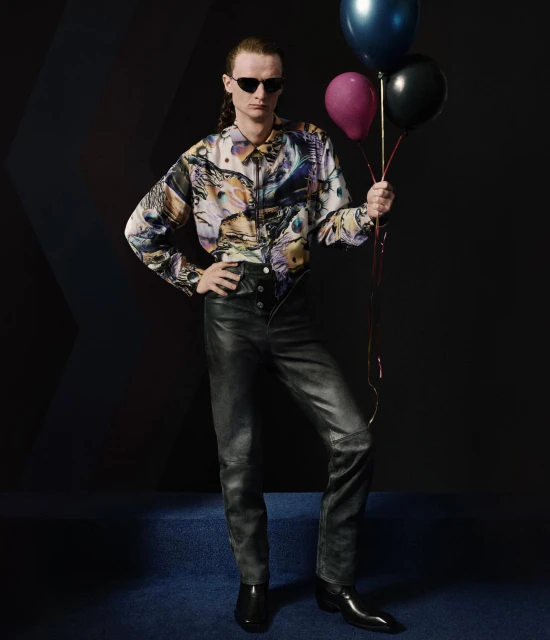 A male model with sunglasses poses in a studio, wearing a printed shirt and leather pants, holding black, blue, and pink balloons.