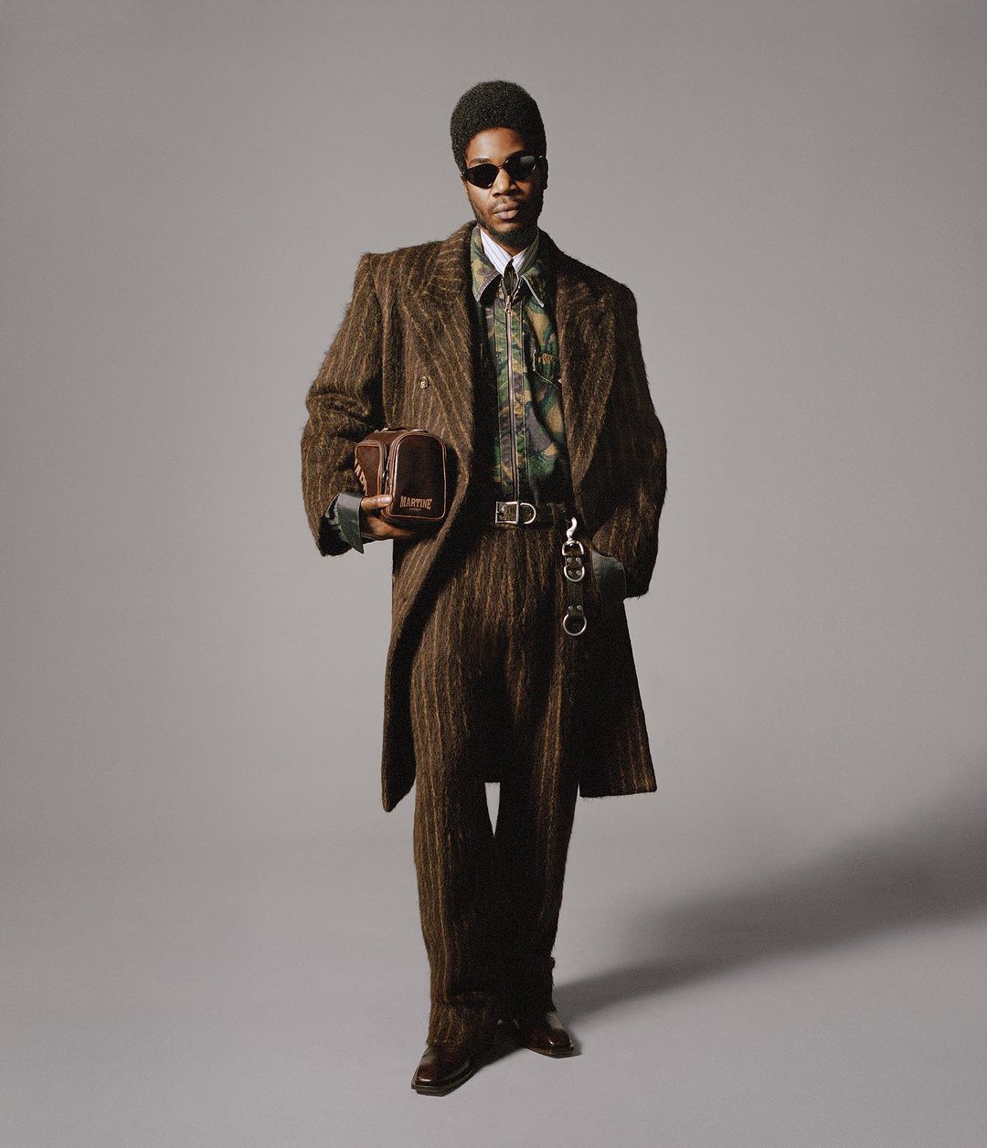 Stylish man in a brown striped suit and sunglasses stands against a gray backdrop, holding a bag, showcasing a fashion-forward aesthetic.
