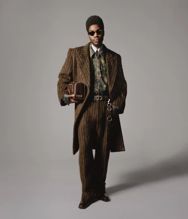 Stylish man in a brown striped suit and sunglasses stands against a gray backdrop, holding a bag, showcasing a fashion-forward aesthetic.