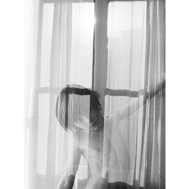 Black and white portrait of a topless person standing in front of a window with sheer curtains, capturing soft, diffused light.
