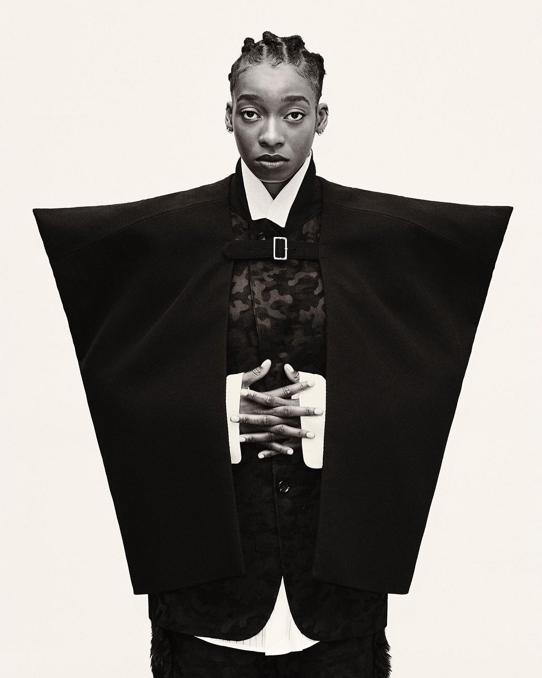 Portrait of a Black adult woman wearing a black kimono-style jacket, with her hands clasped, standing against a white backdrop.