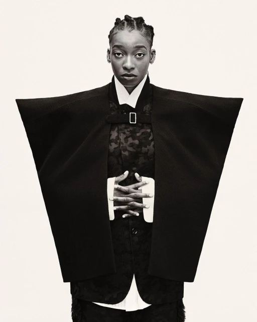 Portrait of a Black adult woman wearing a black kimono-style jacket, with her hands clasped, standing against a white backdrop.