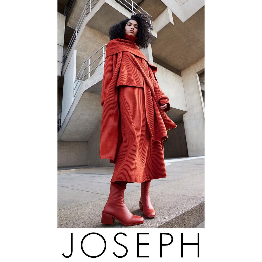 A woman in a head-to-toe orange Joseph knitwear ensemble, featuring a sweater, skirt, and heeled boots, stands against a modern concrete building.