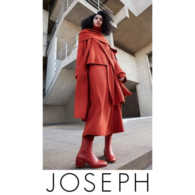 A woman in a head-to-toe orange Joseph knitwear ensemble, featuring a sweater, skirt, and heeled boots, stands against a modern concrete building.