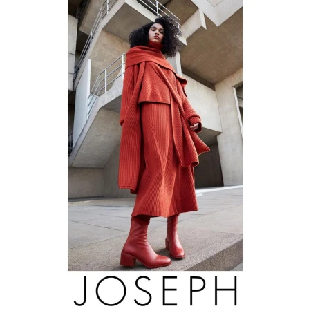 A woman in a head-to-toe orange Joseph knitwear ensemble, featuring a sweater, skirt, and heeled boots, stands against a modern concrete building.