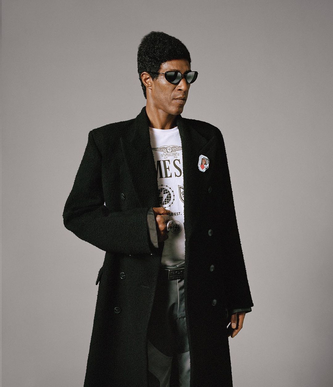 Portrait of a Black man in sunglasses and a long black coat over a white t-shirt, posing in a photo studio setting with a light grey background.