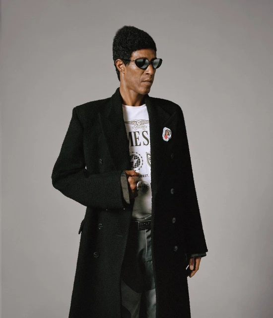 Portrait of a Black man in sunglasses and a long black coat over a white t-shirt, posing in a photo studio setting with a light grey background.