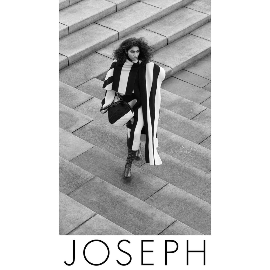 Black and white fashion shot: A woman in a striped poncho and boots walks down a concrete staircase carrying a matching handbag.