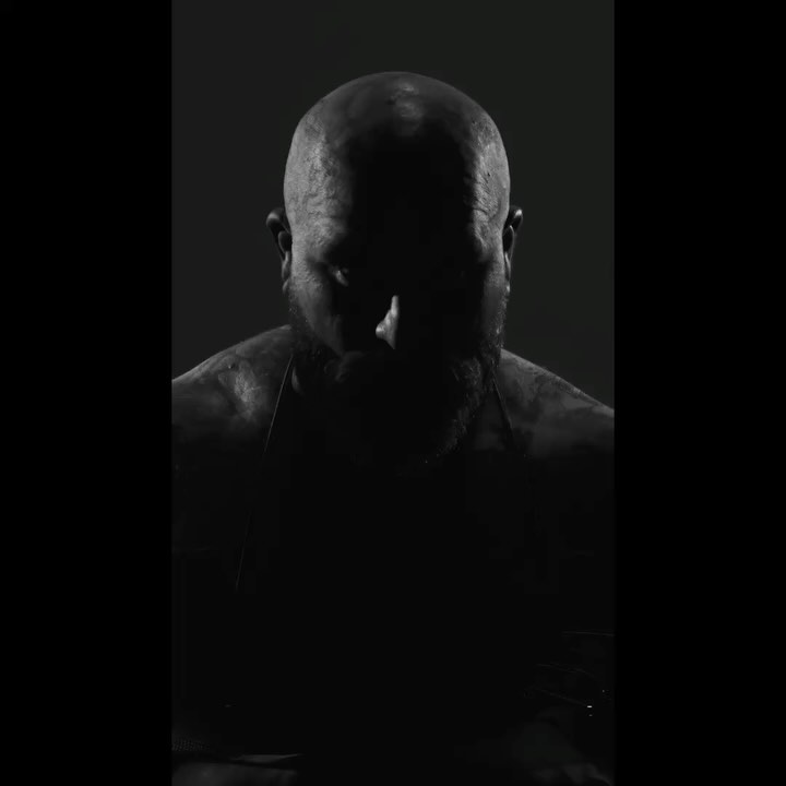 Monochrome portrait of a bald, bearded adult male with a serious expression in dark, low-key lighting against a neutral background.