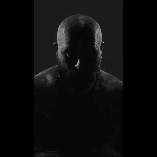 Monochrome portrait of a bald, bearded adult male with a serious expression in dark, low-key lighting against a neutral background.