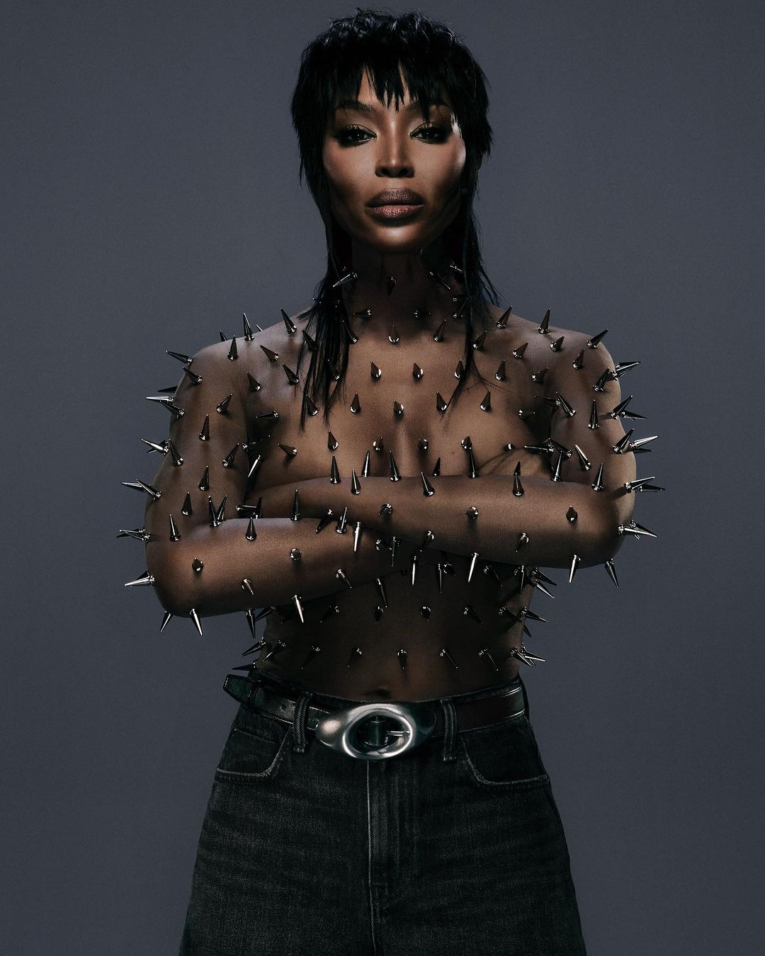 Woman in spike-covered top and dark jeans posing against grey background. Dark hair and makeup. Fashion, beauty, and celebrity concept.