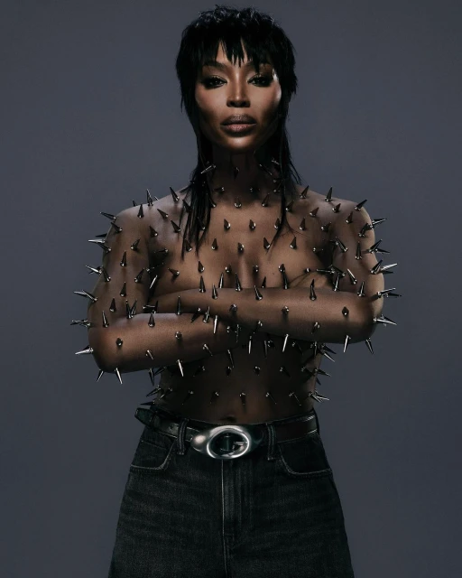 Woman in spike-covered top and dark jeans posing against grey background. Dark hair and makeup. Fashion, beauty, and celebrity concept.