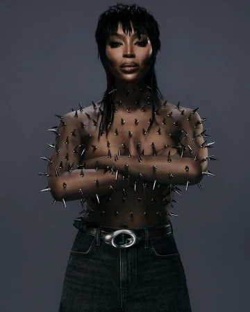 Woman in spike-covered top and dark jeans posing against grey background. Dark hair and makeup. Fashion, beauty, and celebrity concept.