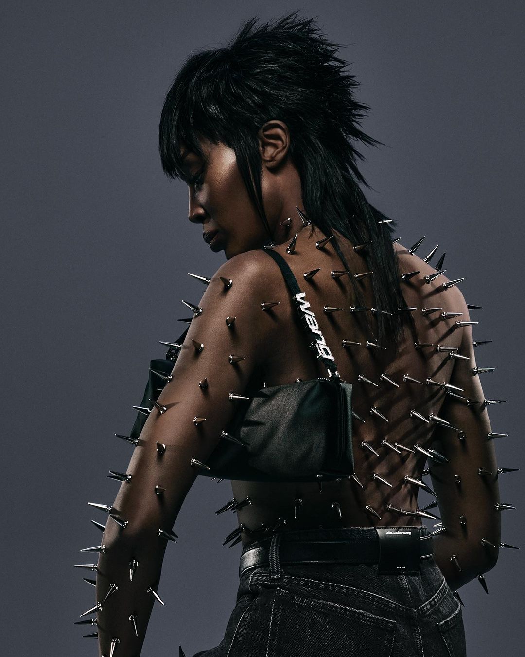 Back view of a Black woman with short dark hair wearing jeans and a studded top, posing in front of a grey background.