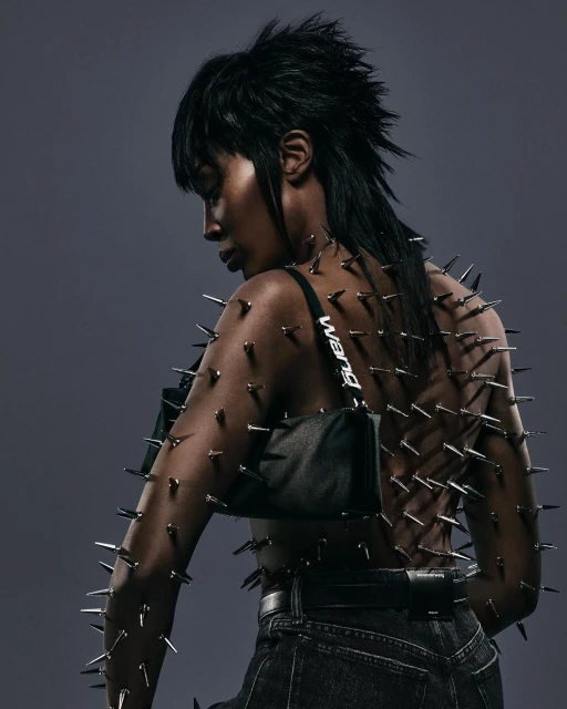 Back view of a Black woman with short dark hair wearing jeans and a studded top, posing in front of a grey background.