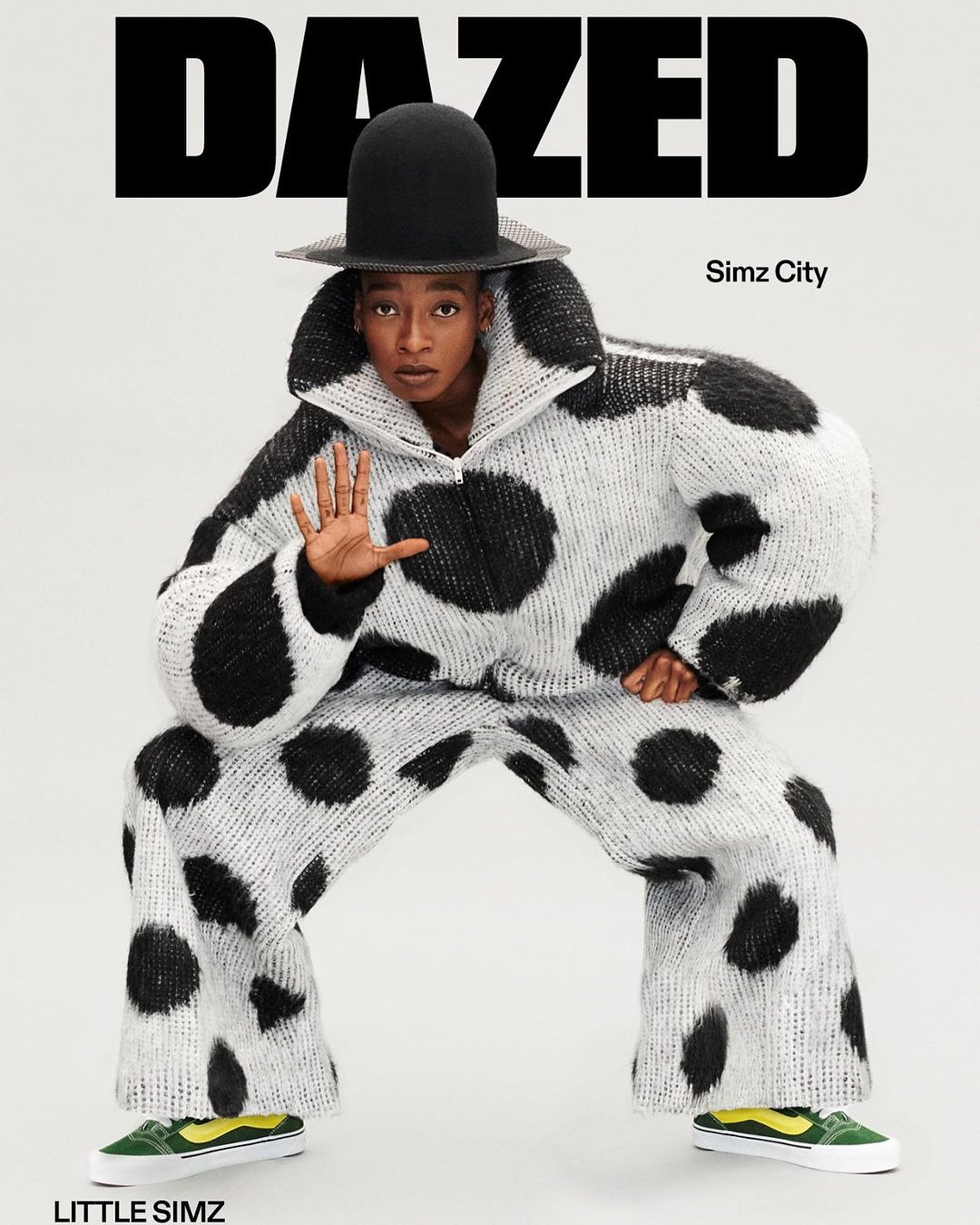 A person wears a black-and-white polka dot jacket and pants, paired with green sneakers and a hat, posing against a white background.