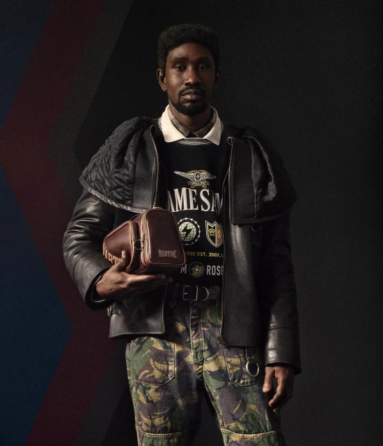 Man in a black leather jacket and camouflage pants holds a leather bag in a studio with a dark background.
