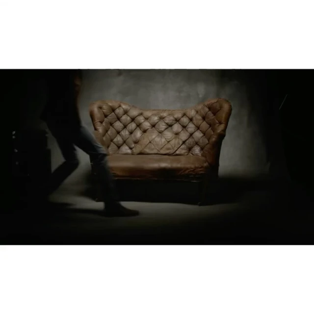 Studio shot of a vintage brown leather tufted couch against a dark grey wall, with a blurry human figure passing by in the shadows.