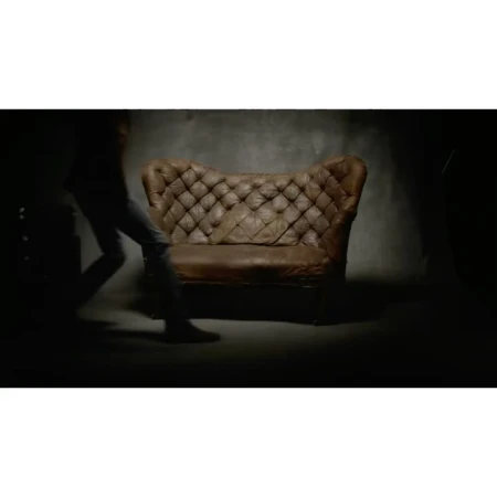 Studio shot of a vintage brown leather tufted couch against a dark grey wall, with a blurry human figure passing by in the shadows.