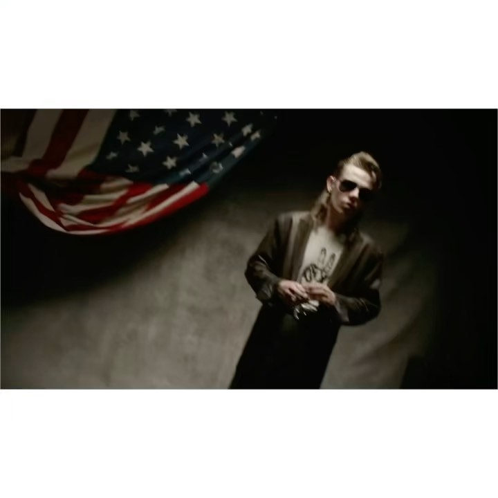 Adult male in sunglasses and coat poses indoors near United States flag. Fashion editorial portrait in studio. Moody lighting.