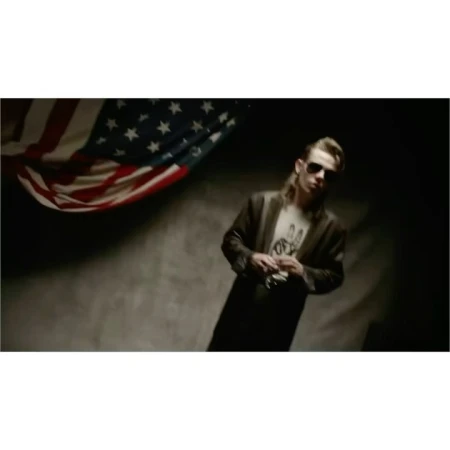 Adult male in sunglasses and coat poses indoors near United States flag. Fashion editorial portrait in studio. Moody lighting.