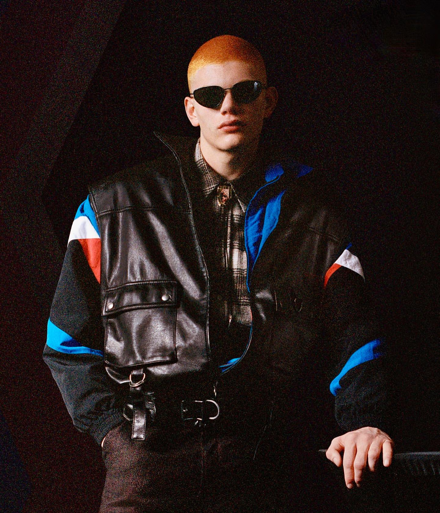 An adult male with a shaved ginger head wearing a dark leather bomber jacket and sunglasses poses in front of a dark background.